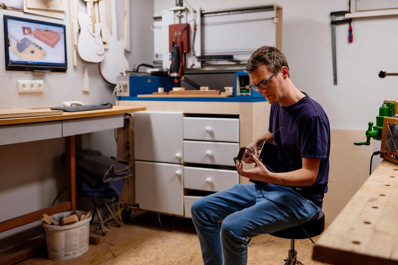 Un homme est assis dans un atelier et fabrique un instrument de musique. À l'arrière-plan, on aperçoit une fraiseuse CNC, un écran et un établi.