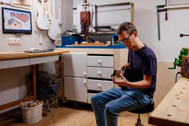 Un homme est assis dans un atelier et fabrique un instrument de musique. À l'arrière-plan, on aperçoit une fraiseuse CNC, un écran et un établi.