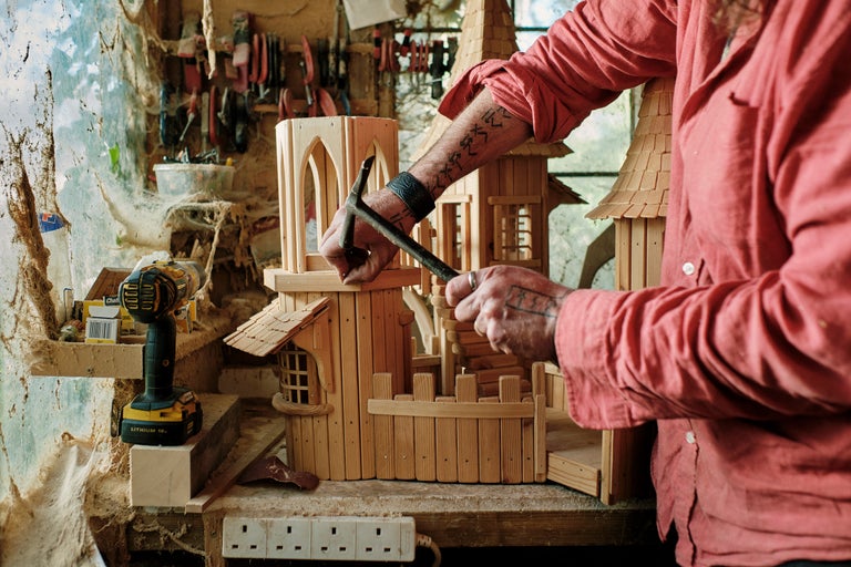 Un homme assemble une maison miniature en bois avec un marteau. Des outils et une perceuse sont visibles à l'arrière-plan.