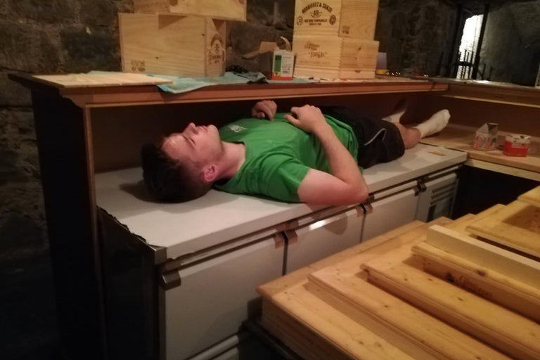 Un homme est allongé sur un établi, entouré de caisses en bois et d'outils, dans un atelier ou une cave.