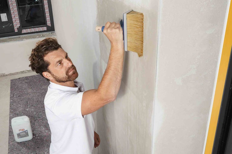 Mann trägt Tiefengrund mit einer Bürste auf eine Wand auf. Im Hintergrund sind ein Fenster und ein Kanister Tiefengrund zu sehen.