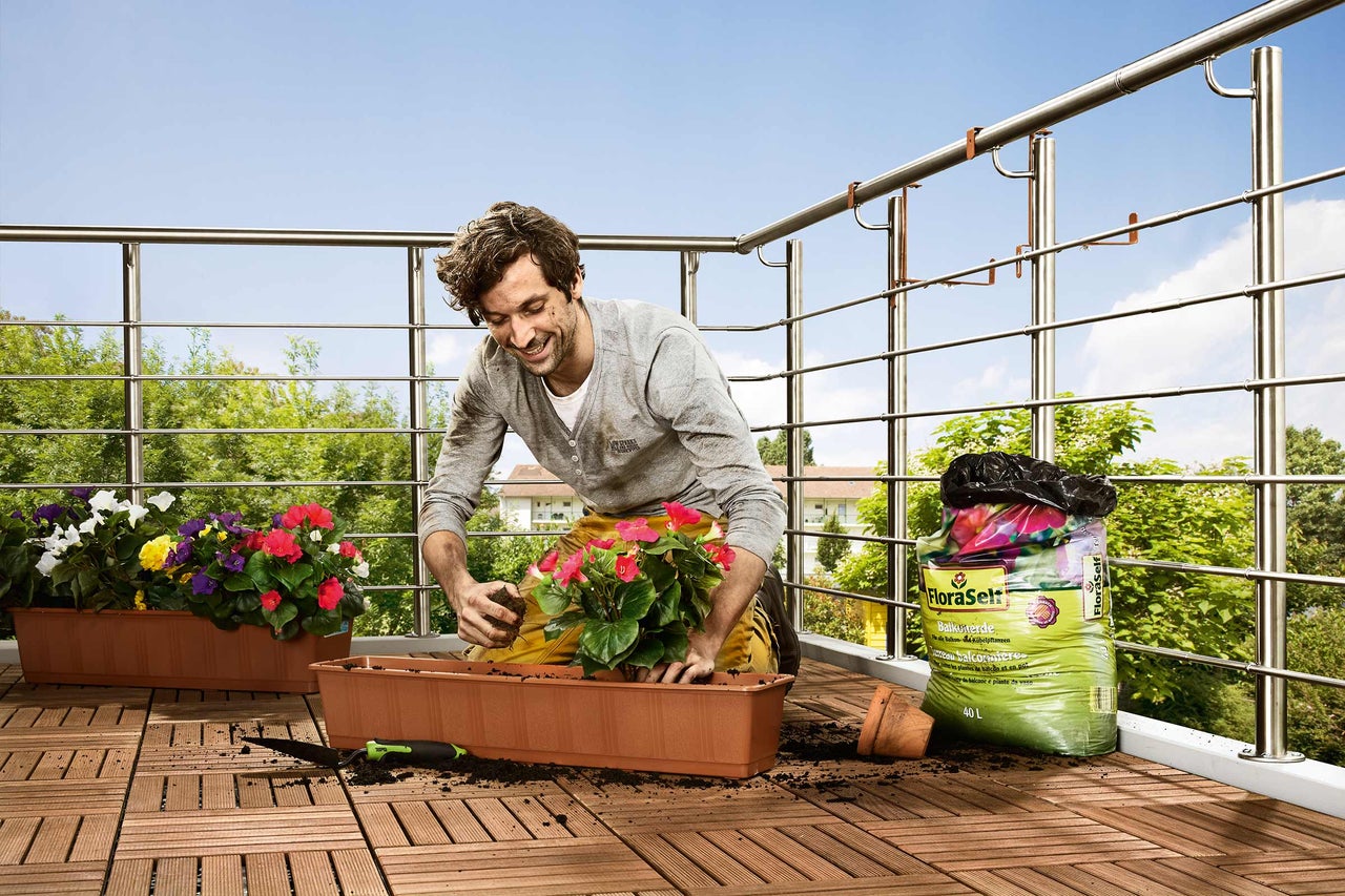 Un homme plante des fleurs sur un balcon avec du terreau dans une jardinière.