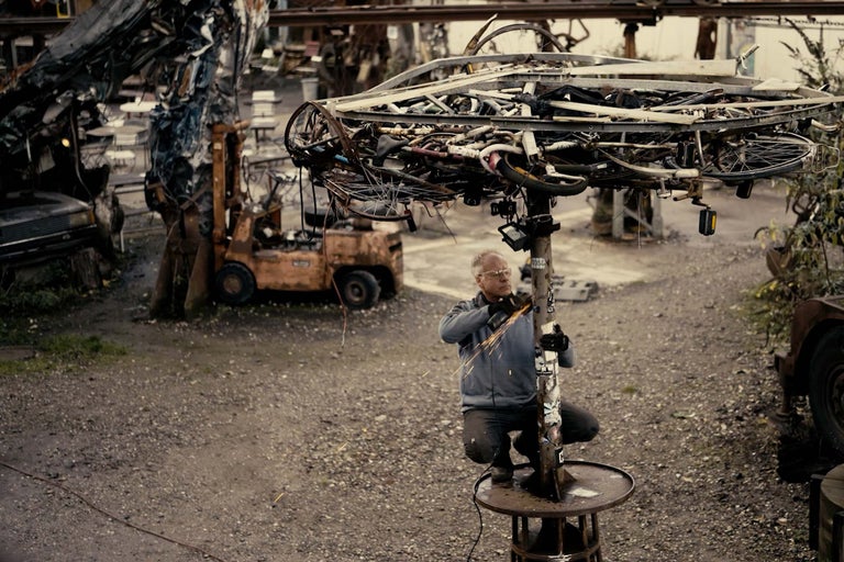 Mann arbeitet mit einem Trennschleifer an einer Metallsäule unter einem Schirm aus Metallschrott.