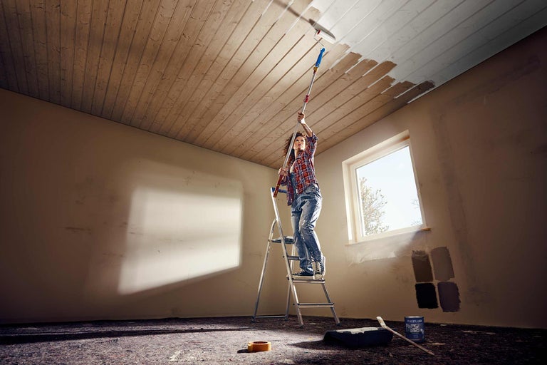 Une femme peint un plafond en bois à partir d'une échelle avec un rouleau à peinture et une perche télescopique. Un bac à peinture, un pinceau et un pot de peinture sont posés sur le sol.