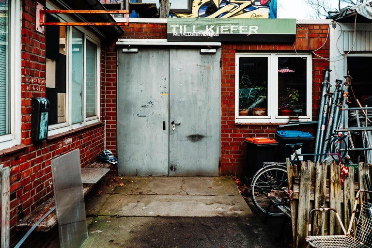 Vue extérieure d'un atelier avec une façade en briques, une porte métallique grise, des fenêtres et un panneau indiquant Till Kiefer Metallgestaltung.