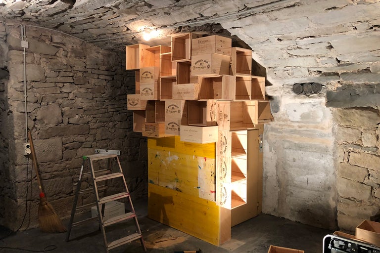 Étagère en caisses de bois dans une cave aux murs en pierre, combinée à des éléments de construction jaunes.