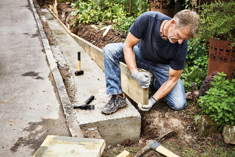Un homme travaille sur une bordure de pelouse en béton avec un coffrage en bois dans un jardin.