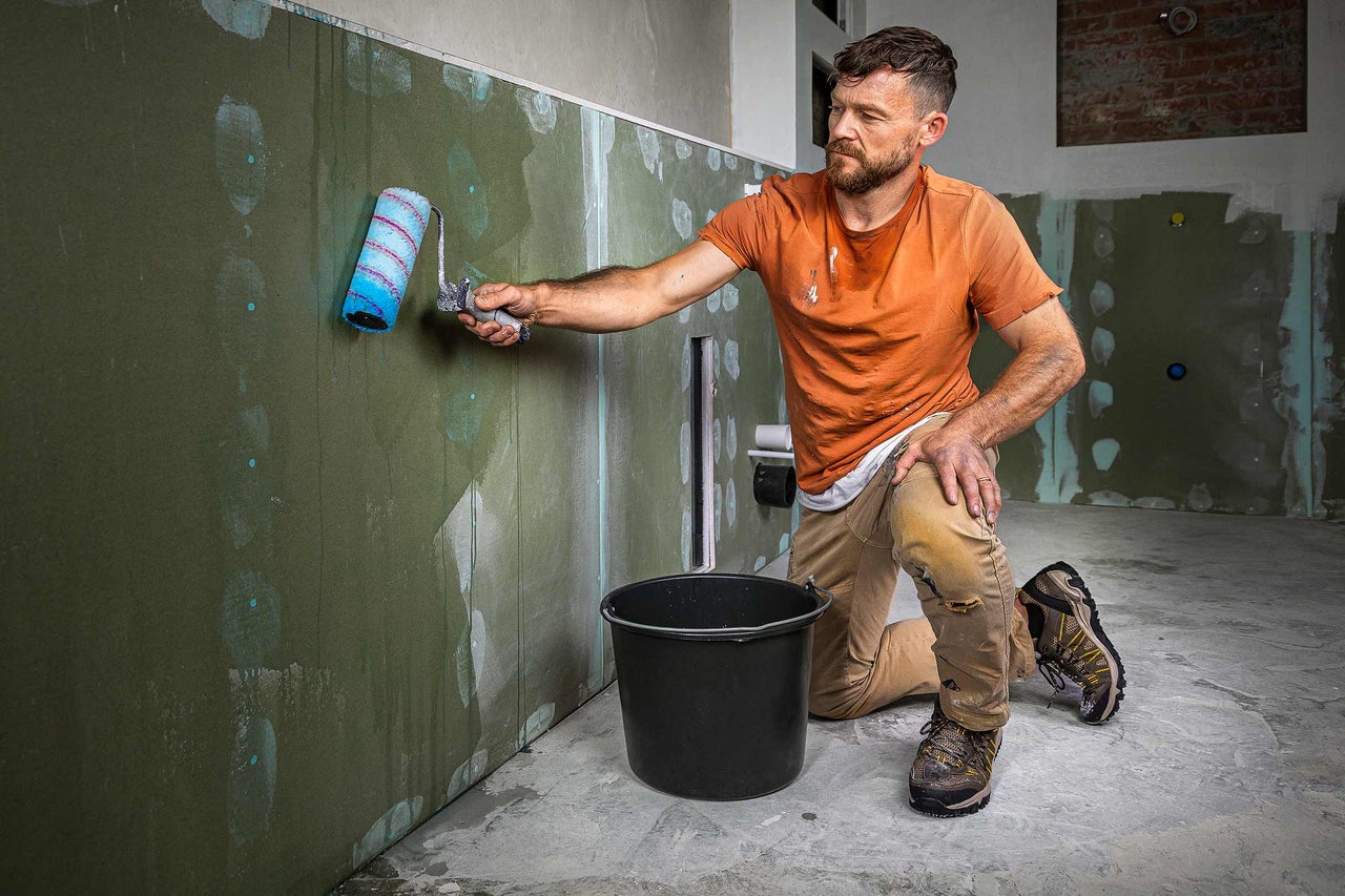 Handwerker streicht Wand mit Farbroller in einem Raum, Eimer steht auf dem Boden