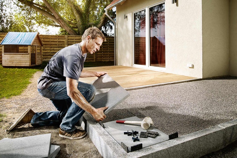 Un homme pose des dalles de terrasse dans le jardin avec des croisillons et un maillet en caoutchouc.