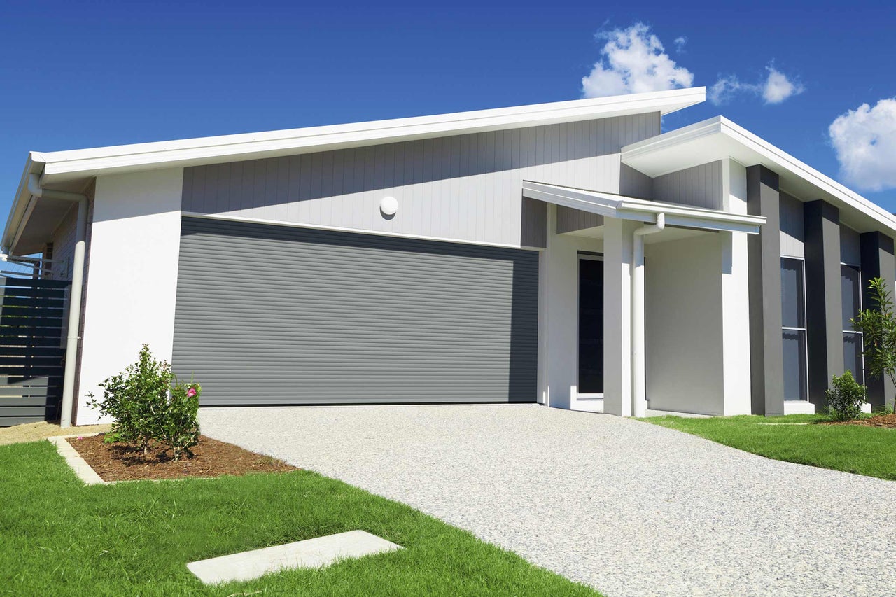 Une maison moderne avec un garage à porte enroulable grise, un chemin pavé et un jardin bien entretenu.