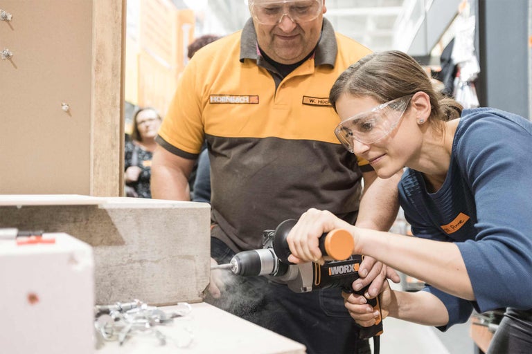 Une femme perce un ouvrage avec une perceuse pendant qu'un homme regarde, tous deux portent des lunettes de protection et la femme un vêtement de travail avec le logo Hornbach.