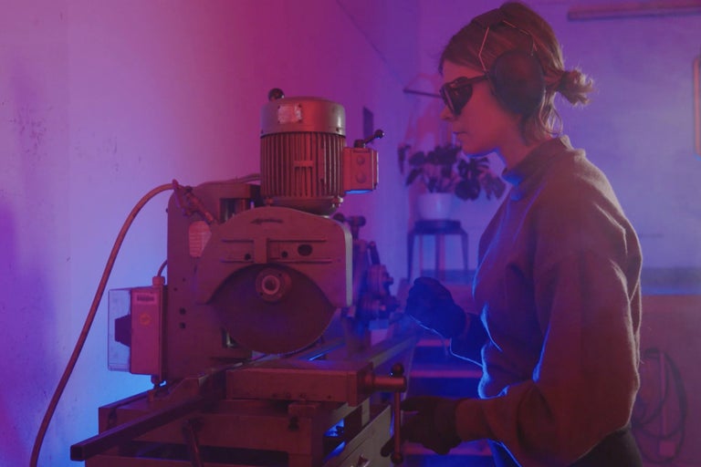 Une femme utilise une scie circulaire à métaux stationnaire avec des lunettes de protection et un casque antibruit.