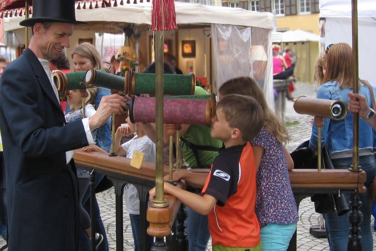 Un homme portant un chapeau haut de forme et des enfants utilisent des télescopes sur un marché.