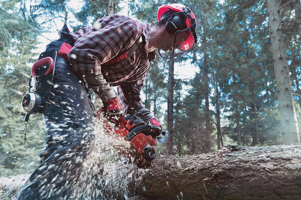 Homme avec casque de protection et protection auditive sciant une bûche à la tronçonneuse dans la forêt