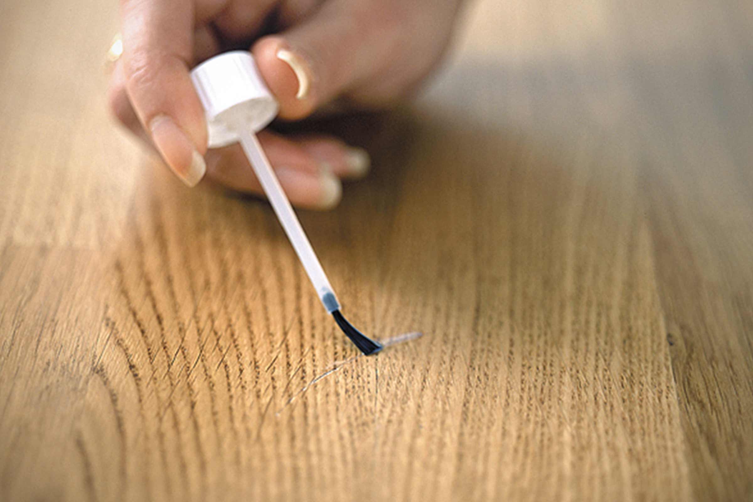 Une personne répare une rayure sur un plancher en bois avec un pinceau de retouche.
