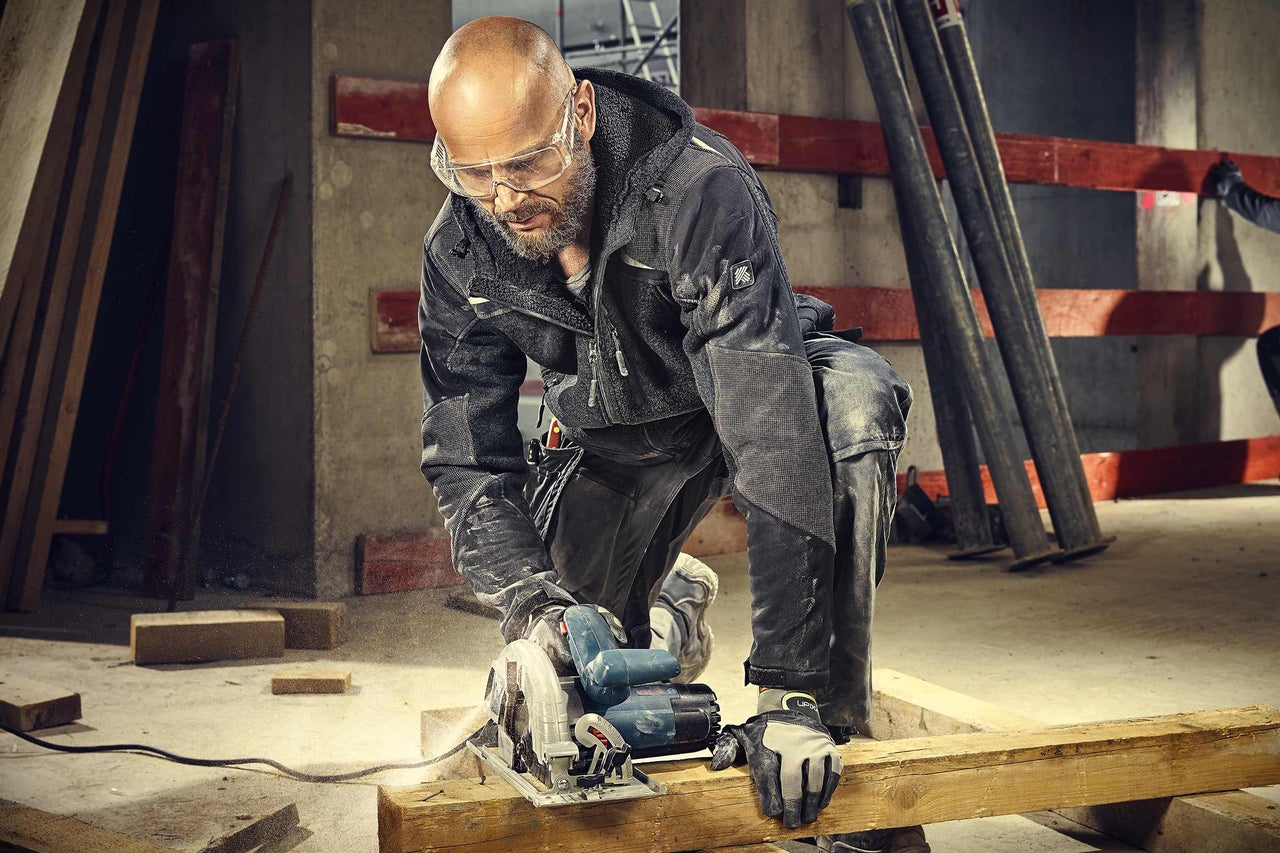 Ein Handwerker sägt mit einer Handkreissäge ein Holzbrett. Der Mann trägt Arbeitskleidung und eine Schutzbrille.