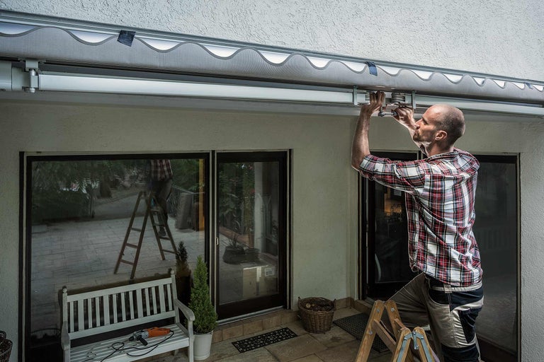 Un homme installe un store banne sur un mur de maison au-dessus d'une porte de terrasse.