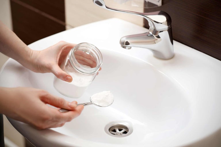 Personne tenant un récipient en verre et une cuillère de poudre nettoyante au-dessus d'un lavabo avec robinet