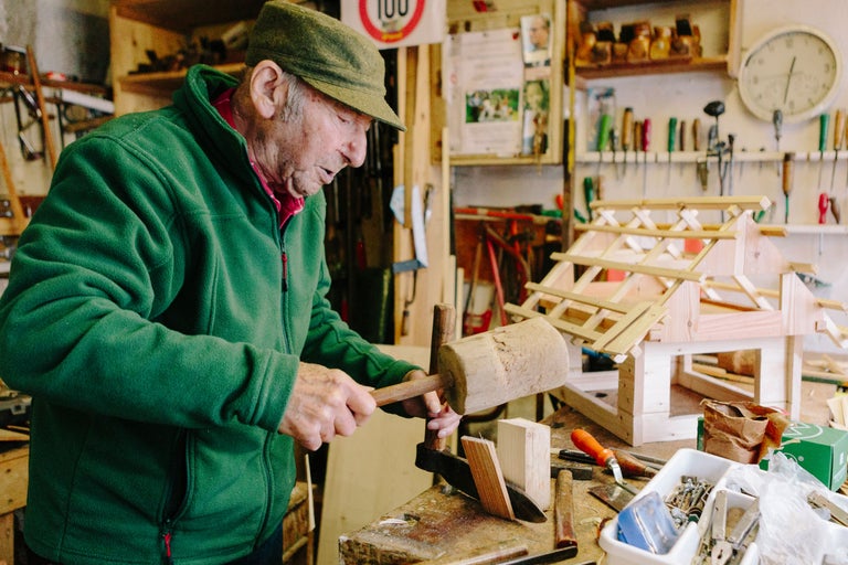 Un homme travaille le bois avec un maillet en bois dans un atelier. Un abri en bois non terminé se trouve à l'arrière-plan.