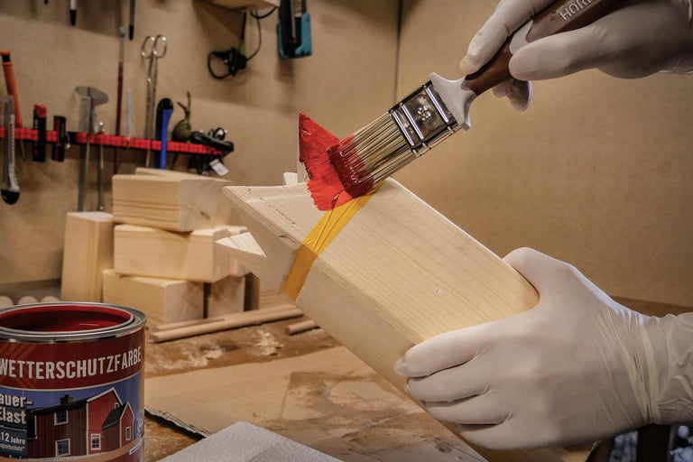 Une personne porte des gants et peint du bois avec un pinceau et de la peinture de protection contre les intempéries rouge.
