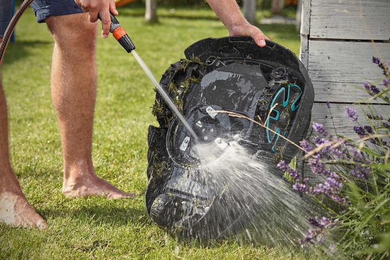 Une personne nettoie un robot de tonte avec un tuyau d'arrosage dans le jardin.