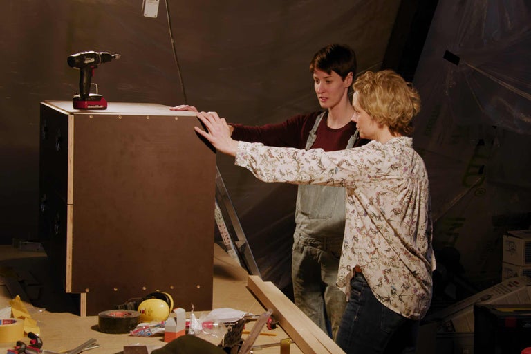 Deux personnes examinent une armoire en bois dans un atelier, avec une perceuse sans fil dessus.