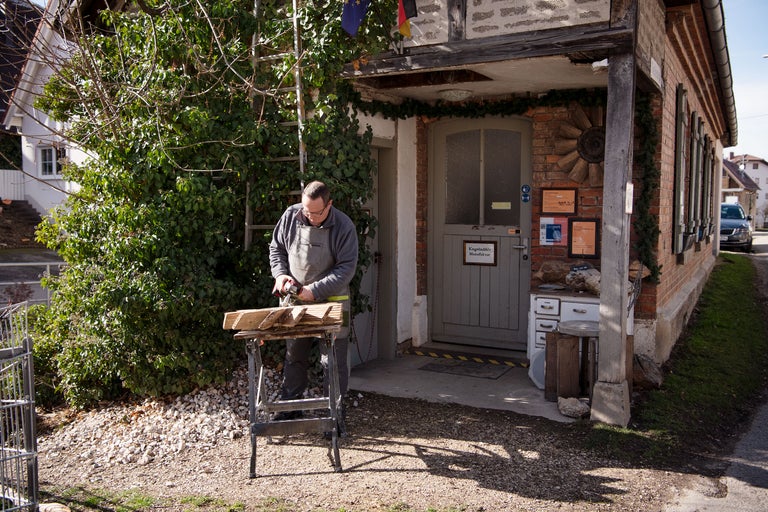 Un artisan travaille à l'extérieur avec une scie sur un chevalet devant une maison.
