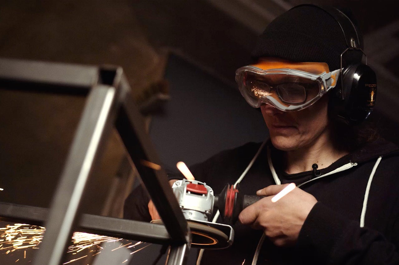 Une personne meule du métal avec une meuleuse angulaire, tandis que des étincelles jaillissent. Elle porte des lunettes de protection et un casque antibruit.