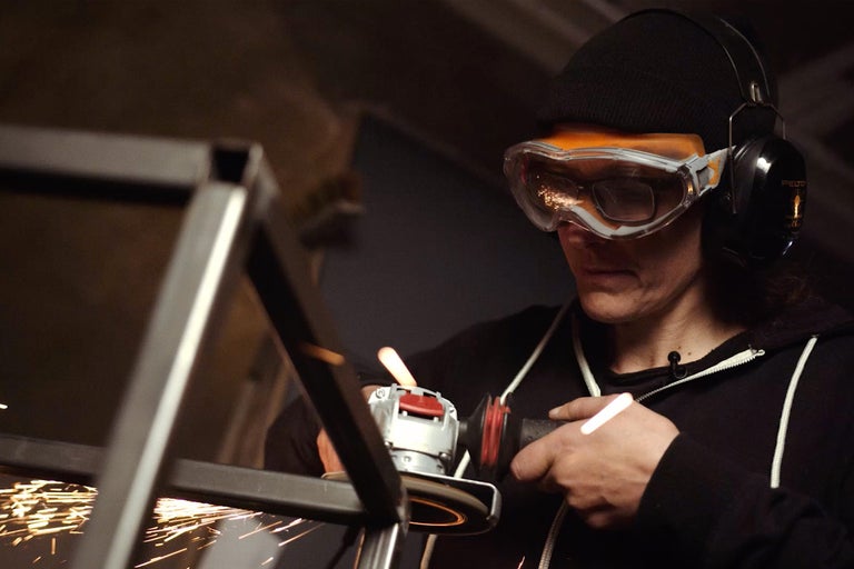 Une personne meule du métal avec une meuleuse angulaire, tandis que des étincelles jaillissent. Elle porte des lunettes de protection et un casque antibruit.