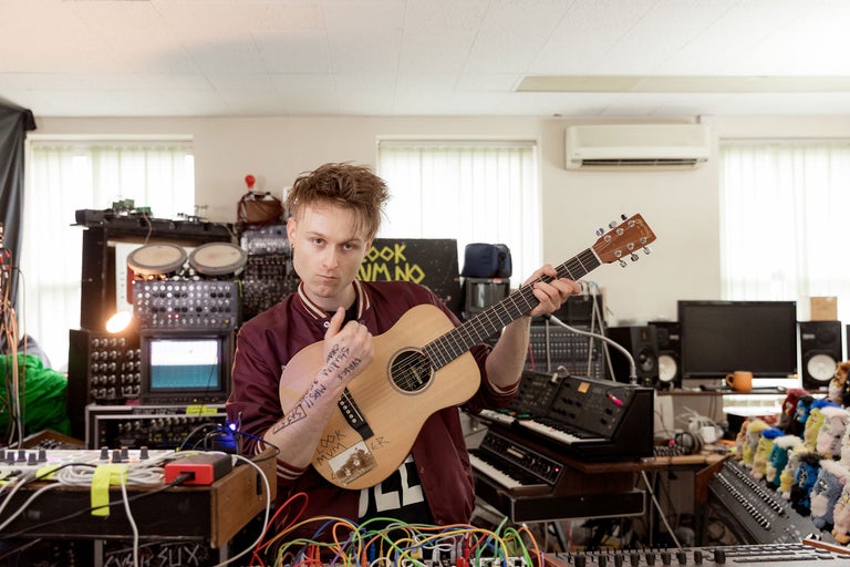 Un homme tient une guitare acoustique dans un studio de musique avec des synthétiseurs et des tables de mixage.