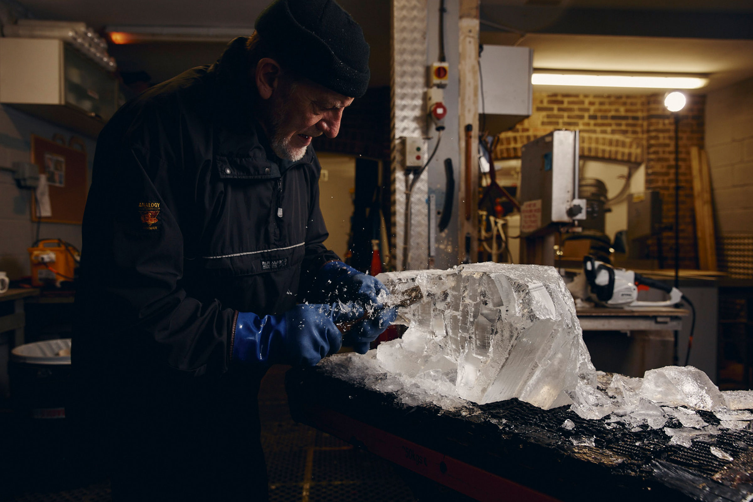Ein Mann mit Handschuhen, der ein Eisblock mit einem Stechbeitel bearbeitet, im Hintergrund Werkzeuge.