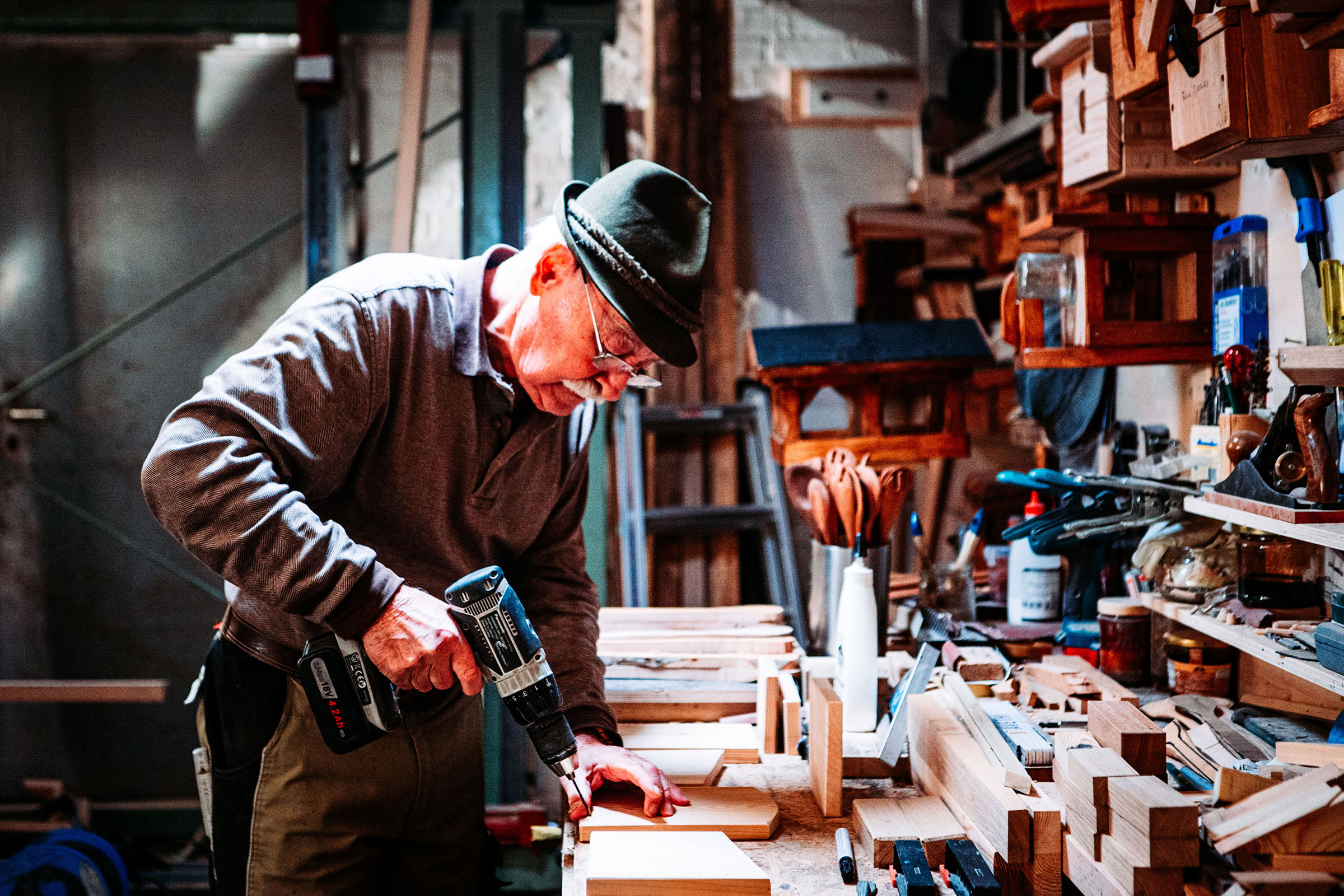 Un homme coiffé d'un chapeau perce du bois avec une visseuse sans fil dans un atelier rempli de nichoirs et d'outils.