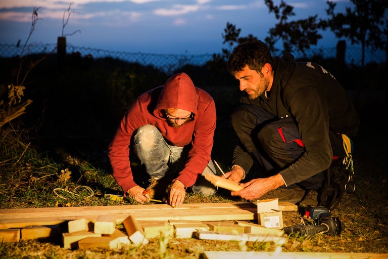 Zwei Personen arbeiten mit Holz im Freien, wobei Werkzeuge wie Massband und Stift verwendet werden.
