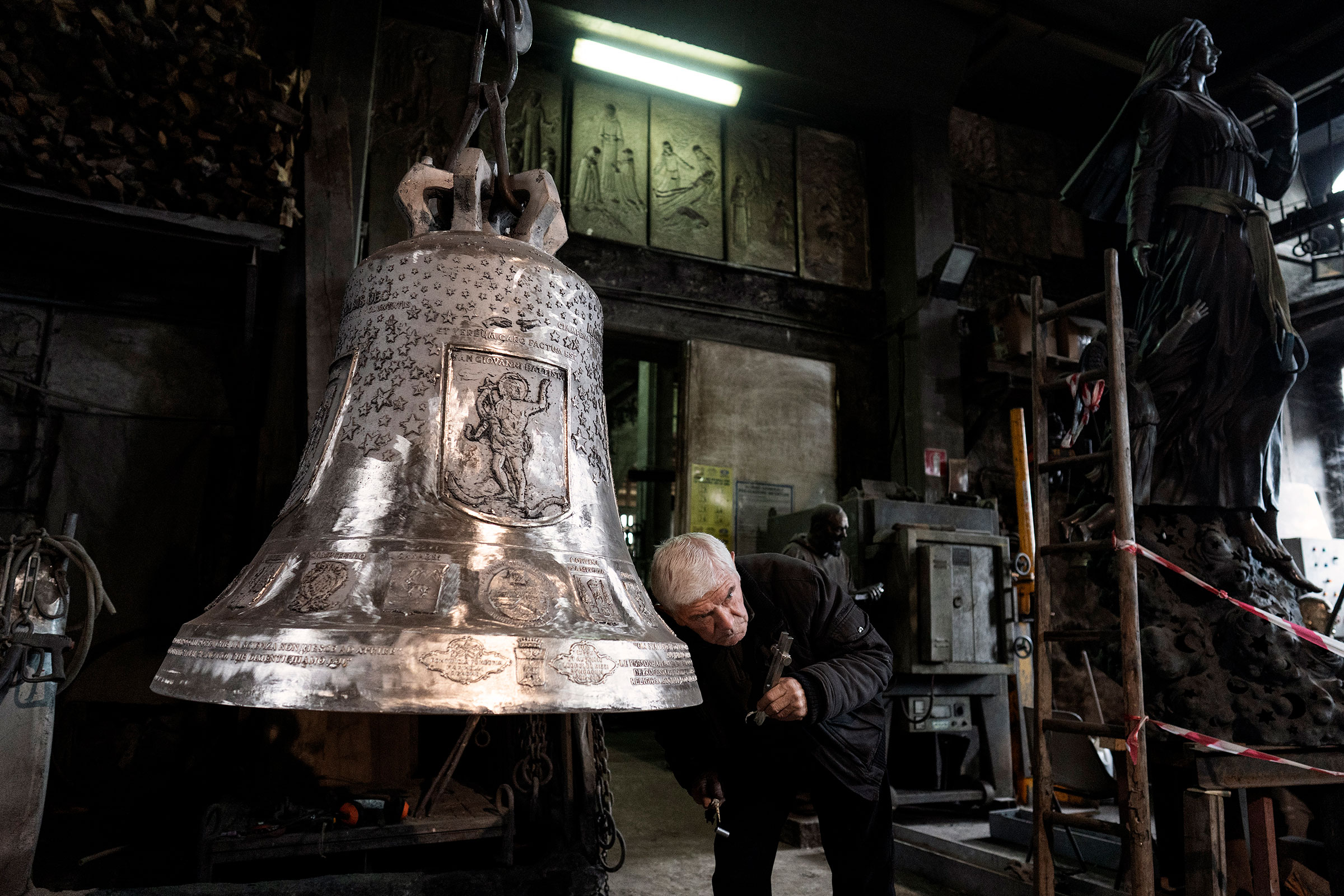 Mann überprüft grosse Glocke in Werkstatt mit Werkzeugen und Statuen