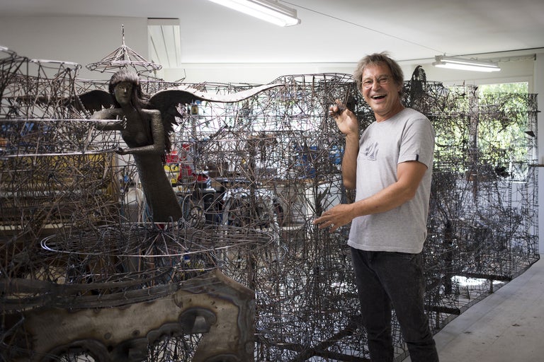 Mann steht neben komplexen Metallskulpturen in einem Werkstattraum.