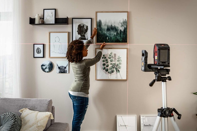 Eine Person hängt mit einem Laser-Nivelliergerät Bilder an einer Wand auf. Verschiedene gerahmte Kunstwerke und Dekorationen sind angeordnet.