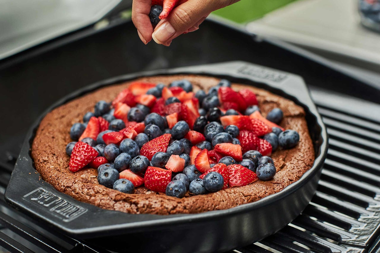 Un gâteau foncé dans un plat à griller avec des fraises et des myrtilles est décoré par une main.