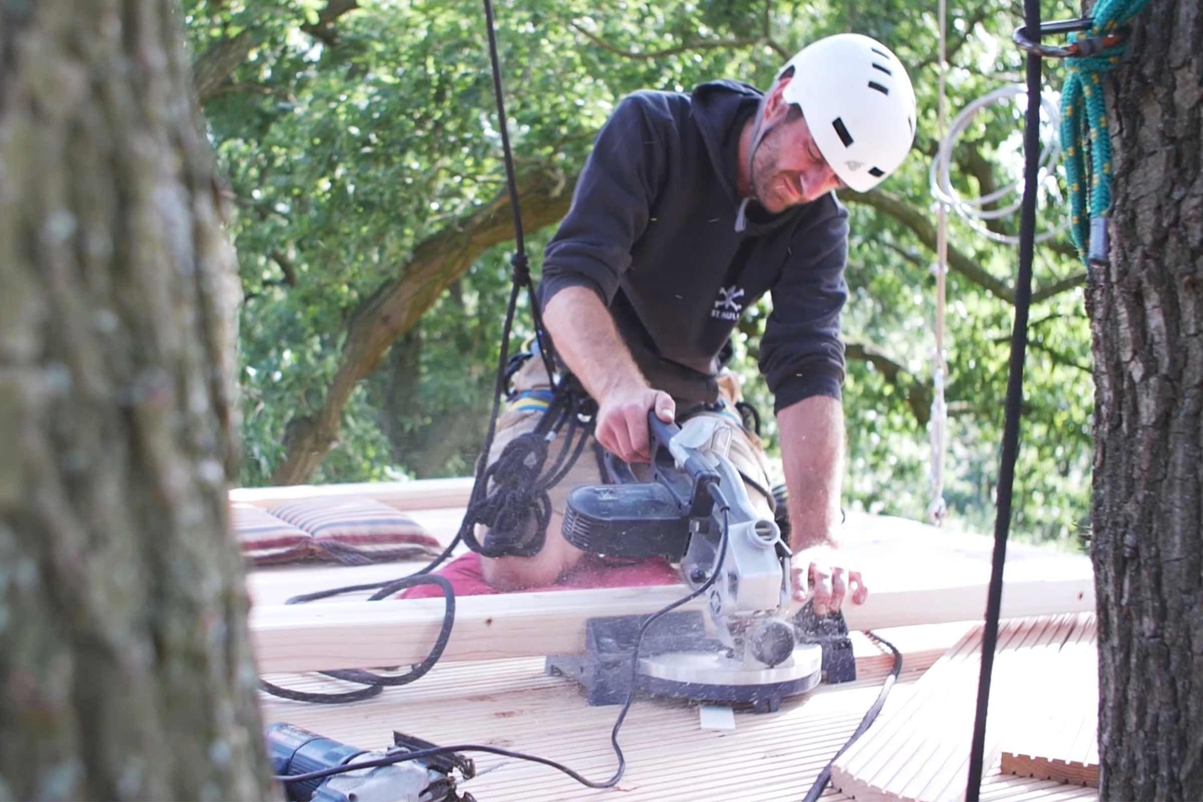 Un homme portant un casque de sécurité coupe du bois avec une scie à onglet lors de la construction d'une cabane dans les arbres.