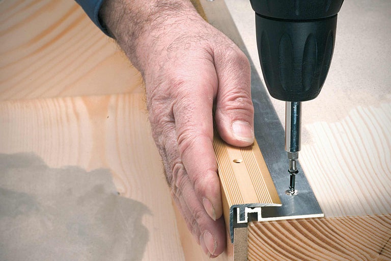 La main fixe le profilé de finition au sol en bois avec une vis et une visseuse sans fil