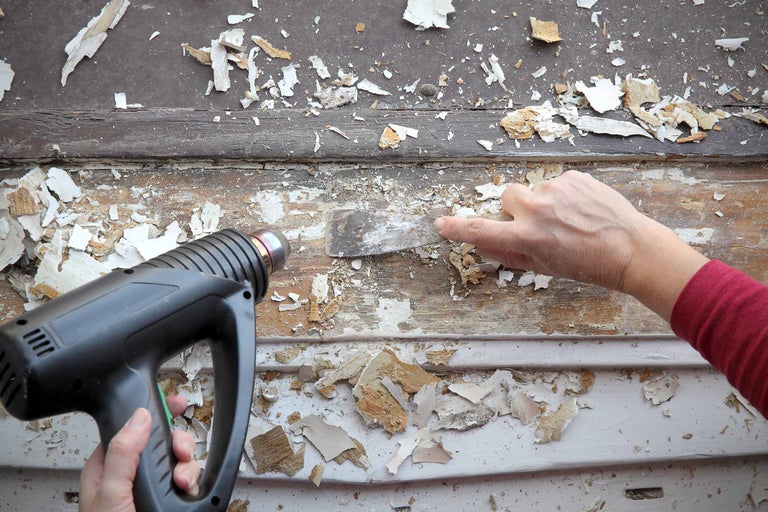 Une personne enlève de vieilles couches de peinture sur du bois à l'aide d'un décapeur thermique et d'un couteau à mastic.