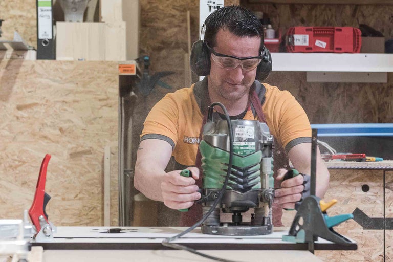 Ein Mann benutzt eine Oberfräse auf einer Holzoberfläche, geschützt mit Brille und Gehörschutz.