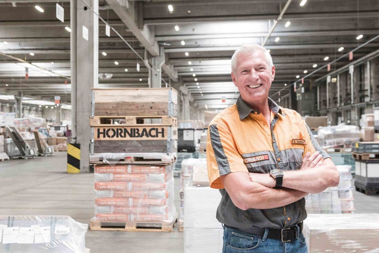 Un collaborateur Hornbach souriant devant un entrepôt avec des palettes et des marchandises.