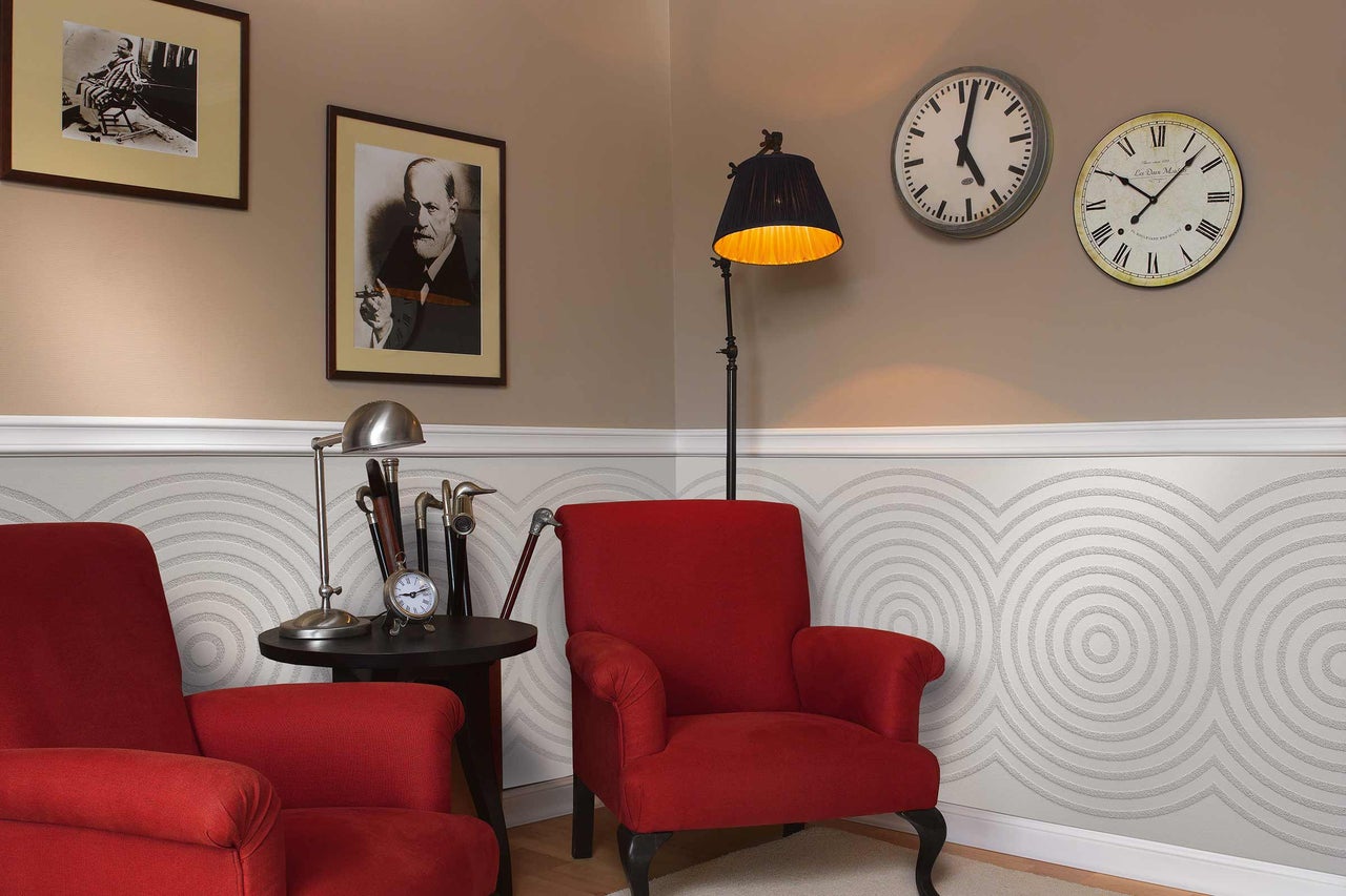 Interieuransicht mit zwei roten Sesseln, Stehlampe, Tischlampe, zwei Wanduhren und dekorativer Wandgestaltung