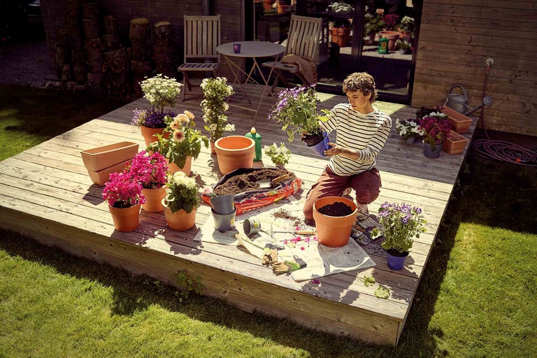 Gartenarbeitsszene auf einer Holzterrasse mit Blumen, Pflanzgefässen, Gartenerde und einer Person.