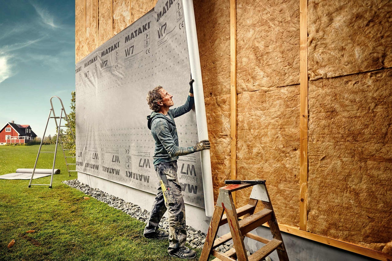 Mann montiert Windschutzplane an einer Hauswand mit Holzrahmen und Wärmedämmung.