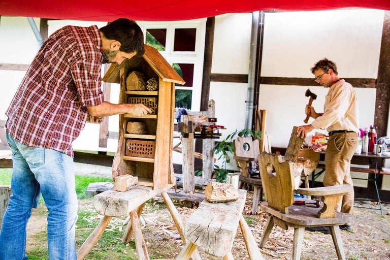 Un homme remplit un hôtel à insectes avec du matériel de nidification, tandis qu'un autre homme hache du bois en arrière-plan