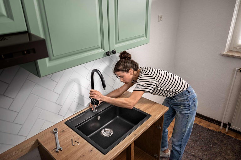 Eine Frau montiert eine schwarze Küchenarmatur an einer dunklen Einbauspüle in einer Küche mit hellen Fronten und Holz Arbeitsplatte.