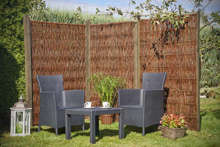 Gartenszene mit Weidenzaun, Rattanstühlen und Tisch auf einer Rasenfläche.