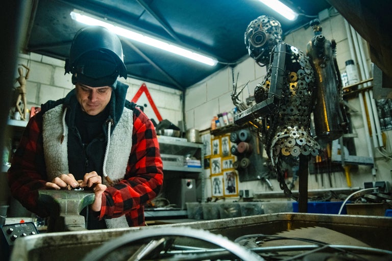 Un homme portant un casque de soudure travaille sur un étau dans un atelier, avec une sculpture en arrière-plan.