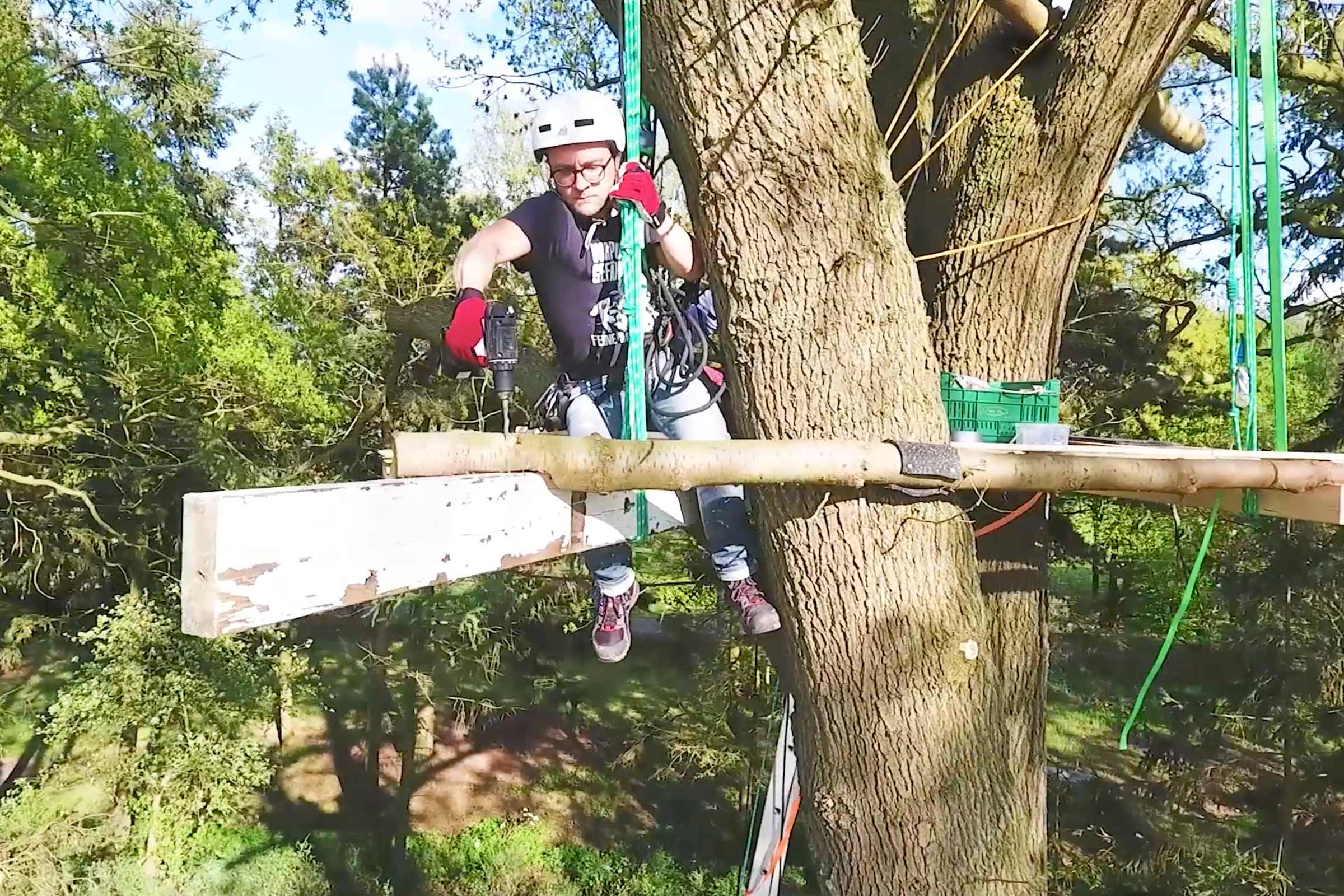 Homme avec casque et équipement de sécurité travaille avec perceuse sur plateforme dans un arbre